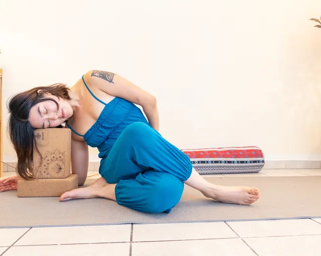 Young Ryu liegt mit geschlossenen Augen auf der Seite, den Kopf auf einem Korkblock, ein Bolster im Hintergrund – Yin Yoga im Studio Sonnenaufgang